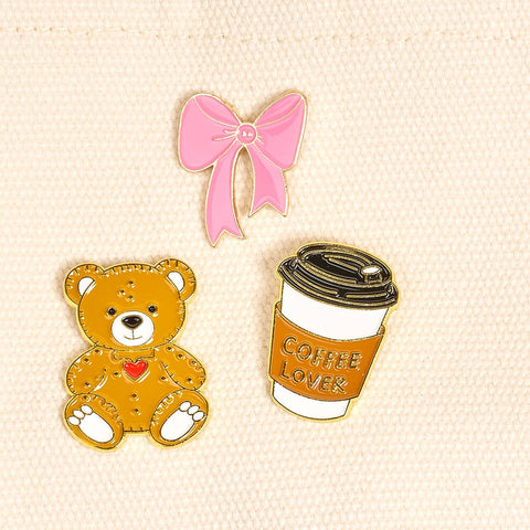 Set of three pins including a teddy bear, coffee cup, and pink bow on a beige background