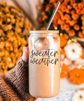 Person holding a glass jar with 'sweater weather' text against an autumn-themed background.