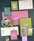 A coordinated stationery set arranged on a dark teal background, including multiple notebooks, a weekly checklist pad, a daily routine notebook, a habit tracker pad, washi tape, a mug, and pens in pastel colors. The set uses several colors in roughly similar prominence: pink, olive green, lavender, white, and dark teal, so the best constrained palette choice is Multicolor.