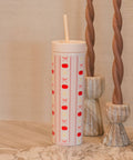 A tall insulated tumbler with a lid and straw, decorated with vertical stripes of  pumpkins, pink bows, and yellow stars on a white background. It is styled on a marble surface next to two twisted brown candles.