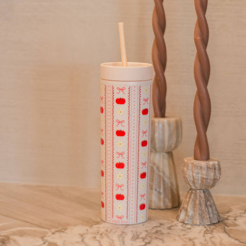 A tall insulated tumbler with a lid and straw, decorated with vertical stripes of  pumpkins, pink bows, and yellow stars on a white background. It is styled on a marble surface next to two twisted brown candles.