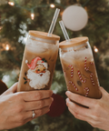 Two hands holding festive iced drinks with Santa Claus design against a Christmas tree background.