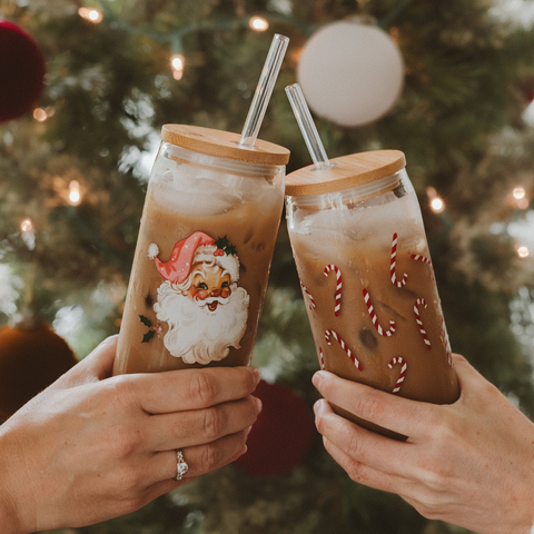 Two hands holding festive iced drinks with Santa Claus design against a Christmas tree background.
