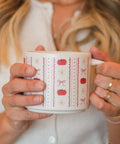 A person holding a white ceramic mug with a red and pink autumn pattern of pumpkins, bows, and zigzags, wearing a white ribbed top and gold rings.