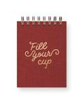 A red spiral-bound notebook with a textured cover and gold foil-style script reading 'Fill Your cup' on the front, shown on a white background.
