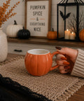 Person holding an orange pumpkin-shaped mug with Halloween decorations in the background.