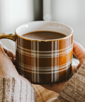 Person holding a plaid mug with coffee in a cozy setting