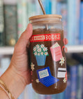 A hand holds a clear glass tumbler with a bamboo lid and metal straw, filled with iced coffee. The tumbler is decorated with illustrations of a potted daisy, a pink ballet dress, a blue laptop with 'AOL' on the screen, a credit card, and a red banner that reads 'BOX & SONS BOOKS'.