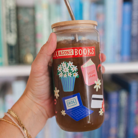 A hand holds a clear glass tumbler with a bamboo lid and metal straw, filled with iced coffee. The tumbler is decorated with illustrations of a potted daisy, a pink ballet dress, a blue laptop with 'AOL' on the screen, a credit card, and a red banner that reads 'BOX & SONS BOOKS'.