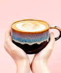 Two hands holding a coffee cup with latte art on a pink background
