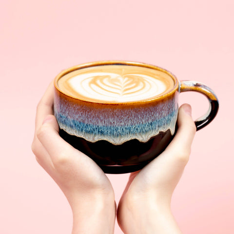 Two hands holding a coffee cup with latte art on a pink background