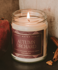 Candle in a jar labeled 'Autumn Orchard' with cinnamon sticks and an apple on a dark surface.