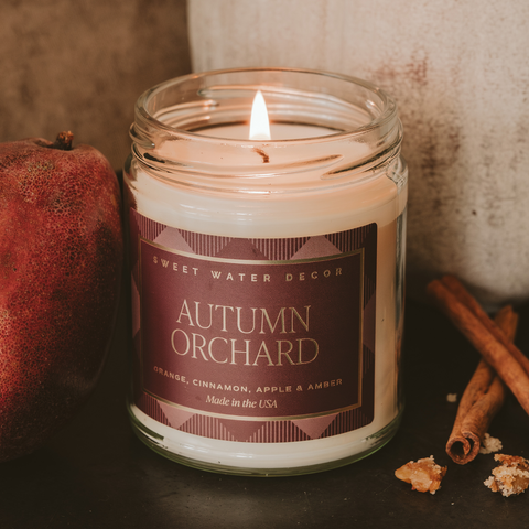 Candle in a jar labeled 'Autumn Orchard' with cinnamon sticks and an apple on a dark surface.
