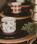 Decorative mug with floral design , surrounded by greenery and a gold Christmas tree.