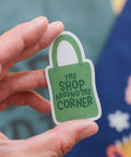 Hand holding a green and white sticker with 'The Shop Around the Corner' text against a blurred background.