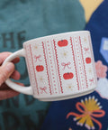 A hand holding a white ceramic mug decorated with a red and white autumn pattern featuring pumpkins, bows, and daisies, set against a colorful, patterned background.