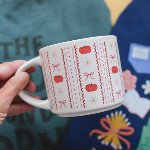 A hand holding a white ceramic mug decorated with a red and white autumn pattern featuring pumpkins, bows, and daisies, set against a colorful, patterned background.
