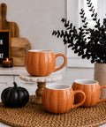 Three orange pumpkin-shaped ceramic mugs with ribbed textures and white interiors, styled on a woven placemat with a small black decorative pumpkin, a candle, a wooden sign reading 'TRICK OR TREAT,' and a potted plant in a bright indoor setting.