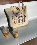 Beige tote bag with 'Let's Go for a Coffee' text on a white bench with two iced coffee cups.