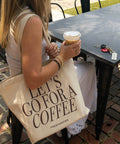 Woman holding a tote bag with 'Let's go for a coffee' text, sitting at an outdoor table.