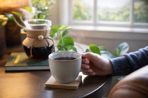 Person holding a mug of coffee with a Chemex in the background on a wooden table.