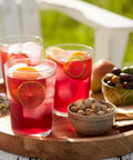 Three glasses of red lemonade with fruit slices on a wooden tray outdoors.