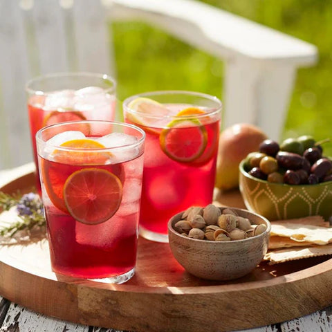 Three glasses of red lemonade with fruit slices on a wooden tray outdoors.