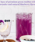 Purple drink in a pitcher with a glass of lemon and blueberries on a white background