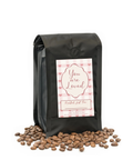Black coffee bag with 'You are Loved' label and scattered coffee beans on a white background