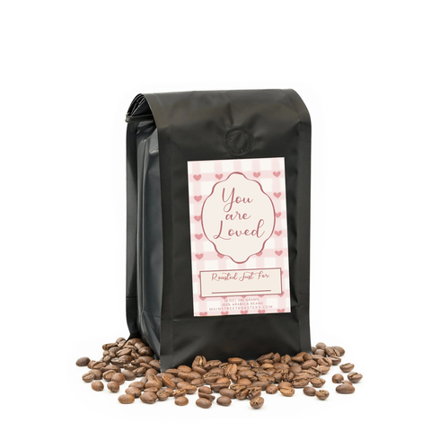 Black coffee bag with 'You are Loved' label and scattered coffee beans on a white background