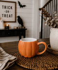 Orange pumpkin mug on a woven mat with Halloween decorations in the background.