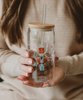 Person holding a glass tumbler with a wooden lid and straw, featuring decorative illustrations.
