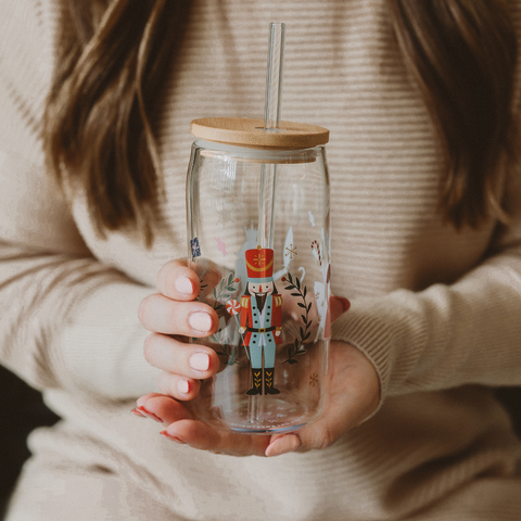 Person holding a glass tumbler with a wooden lid and straw, featuring decorative illustrations.