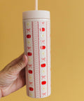 A hand holding a tall insulated tumbler with a lid and straw. The tumbler has a cream background with vertical red and pink decorative stripes, including small pumpkins, bows, and flowers.