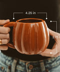 Brown ceramic mug held by hands with measurements indicated, against a blurred background.