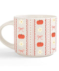 A white ceramic mug decorated with a vertical pattern of orange pumpkins, white daisies, pink bows, and red zig-zag borders on a light cream background.