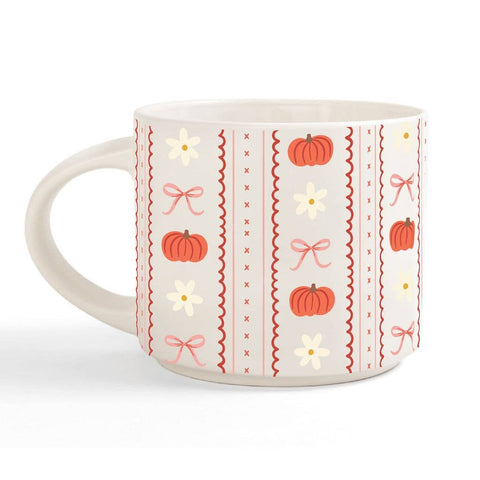 A white ceramic mug decorated with a vertical pattern of orange pumpkins, white daisies, pink bows, and red zig-zag borders on a light cream background.