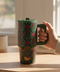 A hand holding a dark green travel tumbler with a festive red and green holiday pattern, set on a wooden table in a sunlit room.