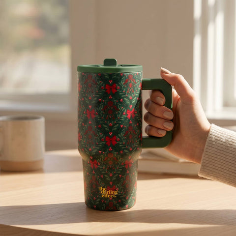 A hand holding a dark green travel tumbler with a festive red and green holiday pattern, set on a wooden table in a sunlit room.