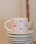 A white ceramic mug with a red and pink autumn-themed pattern of pumpkins, bows, and zigzags, sitting on a stack of white plates. The background features a warm beige wall and decorative twisted wooden objects.