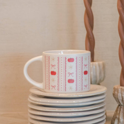 A white ceramic mug with a red and pink autumn-themed pattern of pumpkins, bows, and zigzags, sitting on a stack of white plates. The background features a warm beige wall and decorative twisted wooden objects.