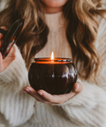 Person holding a lit candle in a dark glass holder with a blurred background