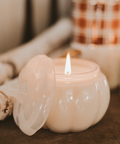 White pumpkin-shaped candle with a lit flame on a brown surface.