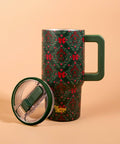 A dark green insulated tumbler with a festive red and green holly pattern, shown with its matching green handle and clear lid resting beside it on a warm peach background. The brand name 'the sailing effect.