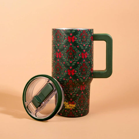 A dark green insulated tumbler with a festive red and green holly pattern, shown with its matching green handle and clear lid resting beside it on a warm peach background. The brand name 'the sailing effect.