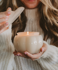 Person holding a lit candle in a white ceramic holder, wearing a beige sweater.