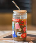 A clear glass jar tumbler with a bamboo lid and metal straw, filled with a brown beverage. The glass features colorful decorative stickers including a red 'BOOKS' label, a pink book sticker, and a red bow sticker, resting on a tray in a warm, blurred indoor setting.