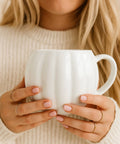 A close-up lifestyle shot of a woman in a cream knit sweater holding a white pumpkin shaped ceramic mug with both hands. She is wearing delicate gold rings.