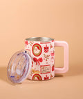 A pink handled mug with a clear lid, decorated with a red and gold Christmas pattern including Santa Claus, bows, and ornaments, set against a solid beige background.
