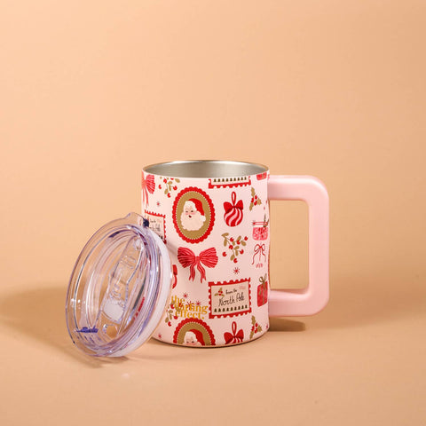 A pink handled mug with a clear lid, decorated with a red and gold Christmas pattern including Santa Claus, bows, and ornaments, set against a solid beige background.